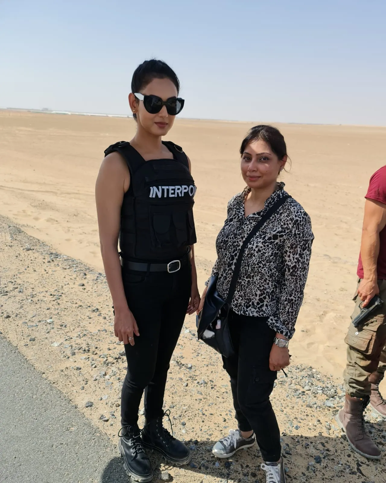 Two women standing on desert sand, one in sunglasses and a black vest and one in a patterned jacket