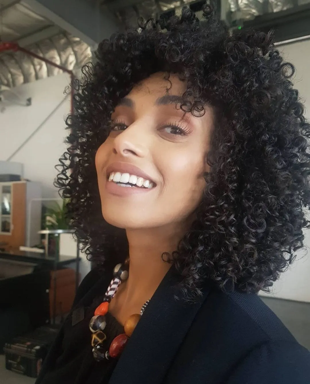 Smiling woman with voluminous curly dark hair in a black jacket and beaded necklace