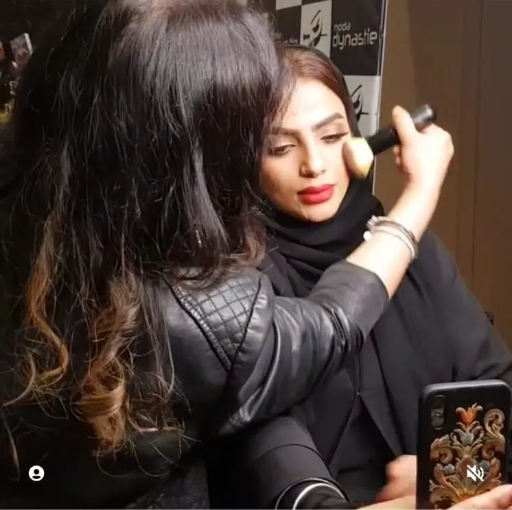 Makeup artist in a black jacket painting the face of a woman in a black headscarf