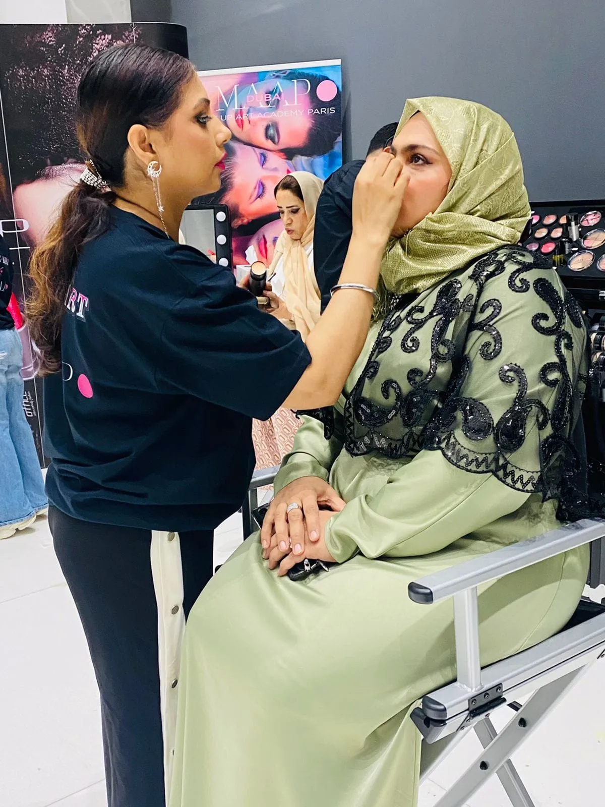 Makeup artist in a dark top applying makeup to a woman in a sage-green headscarf and lace abaya, seen from the side