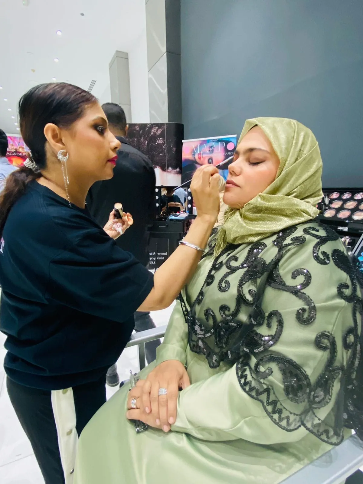 Makeup artist in black working on the eye makeup of a woman in a sage-green headscarf and lace abaya