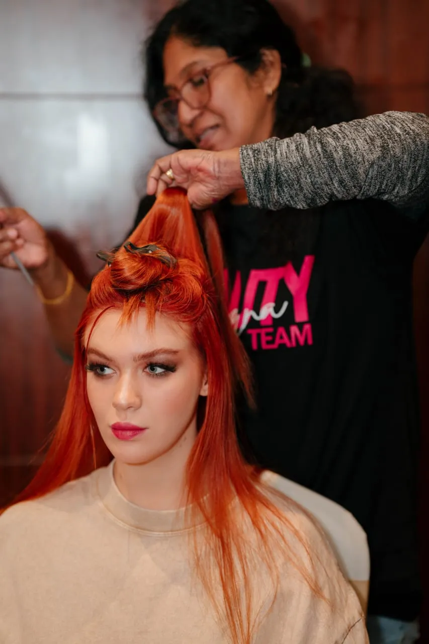 Hair stylist working on the long orange-red hair of a woman in a beige top