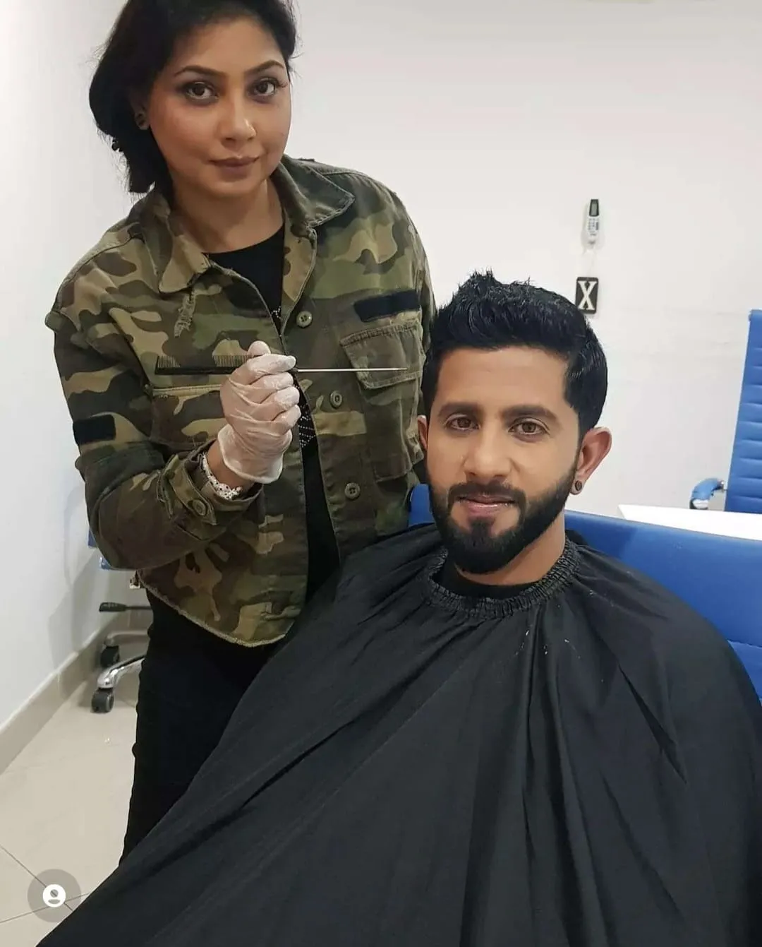 Artist in a camouflage jacket working on a bearded man seated in a black cape