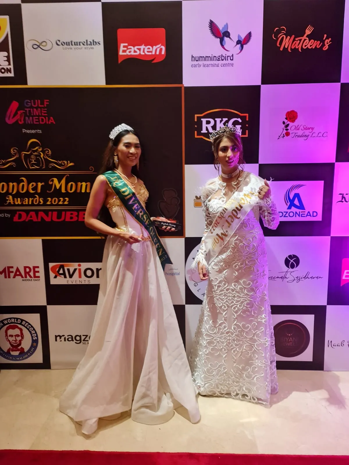 Two women in long gowns with sashes standing apart in front of an event backdrop, full length