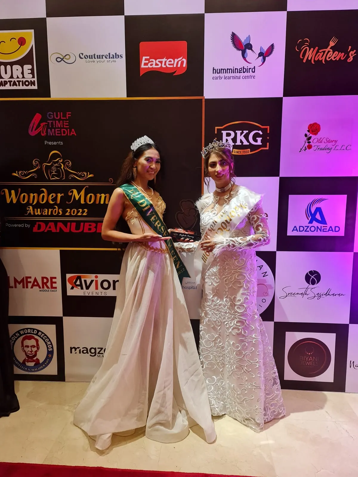 Two women in long gowns with sashes and tiaras standing close together in front of a sponsor backdrop