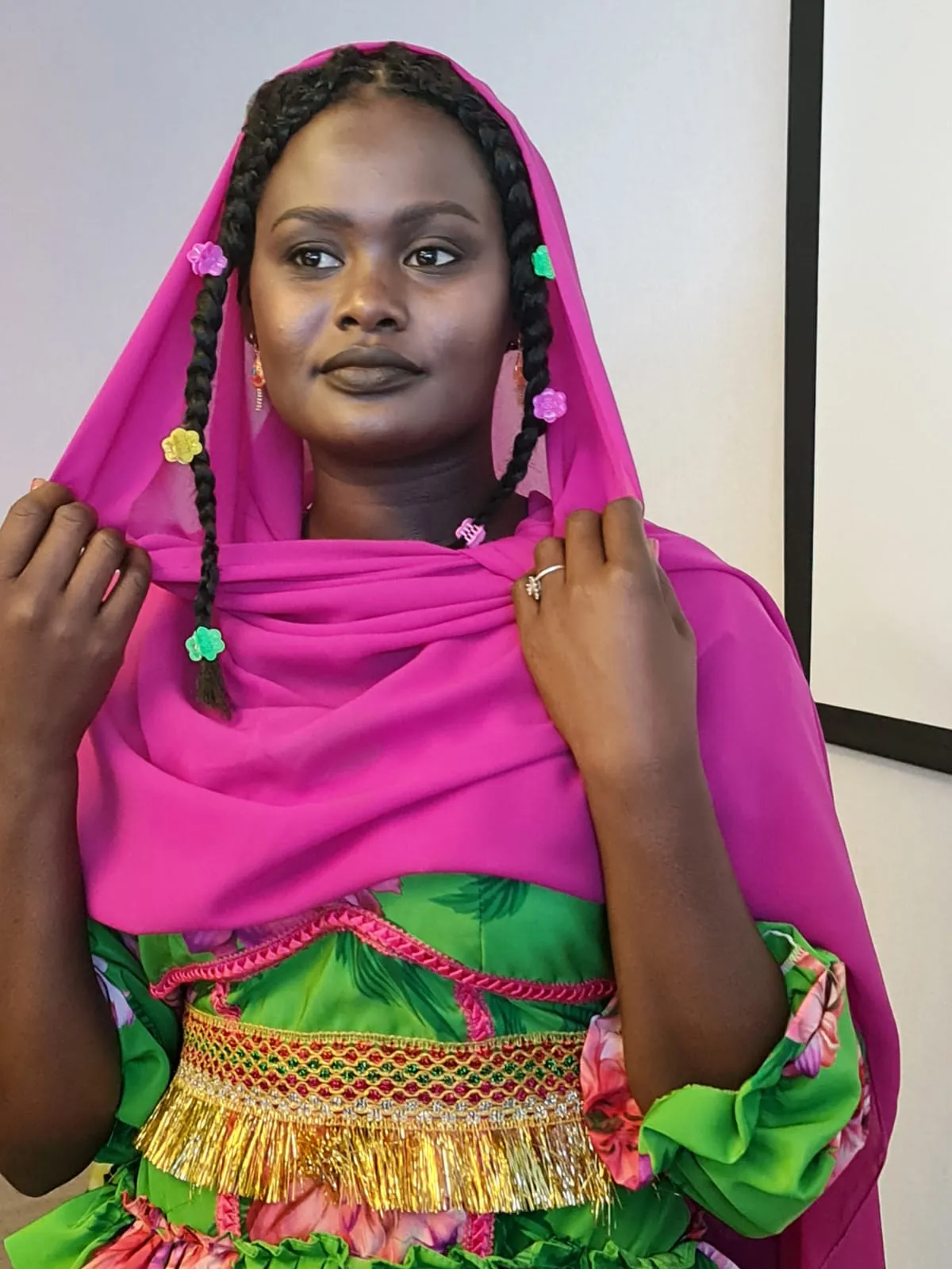 Woman in a bright pink and green patterned outfit with a pink headscarf and beaded braids