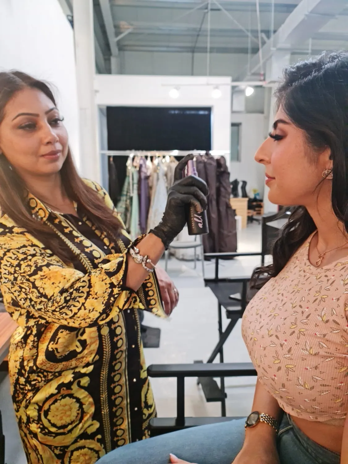 Makeup artist in a gold baroque-print jacket preparing a model's look beside a backstage clothes rail