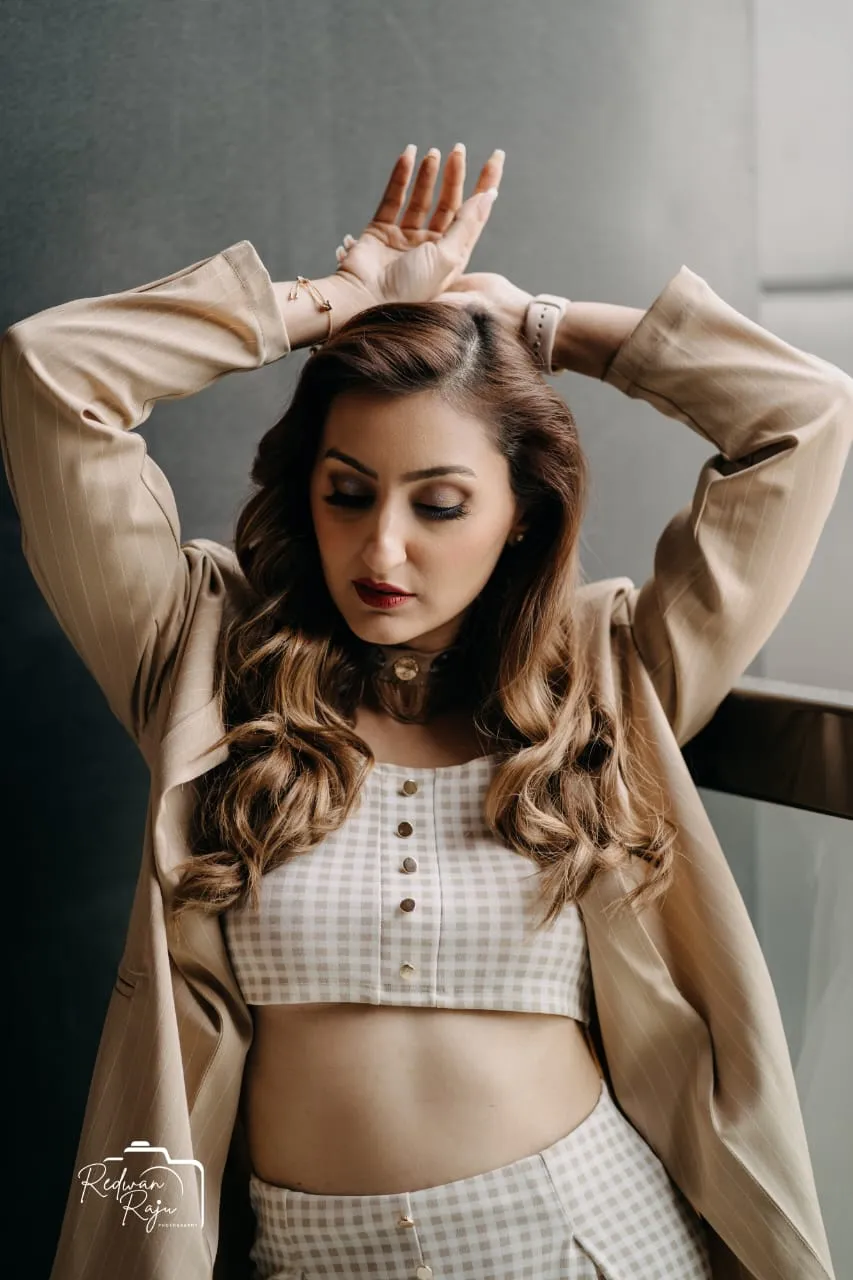 Woman in a beige jacket and cream crop top with wavy hair and her arms raised above her head