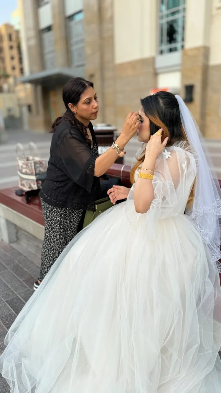 Makeup artist adjusting the hair and veil of a bride in a white gown in front of a building