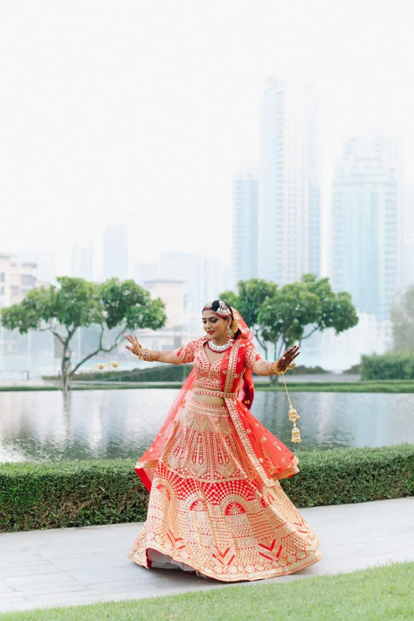 Woman in a red embellished lehenga twirling in a park beside a lake with a city skyline behind