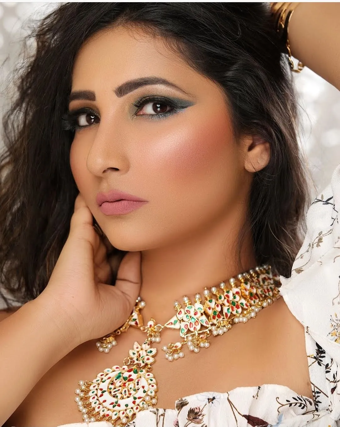 Woman with dark wavy hair, smoky green eye makeup and a gold choker, one hand at her neck