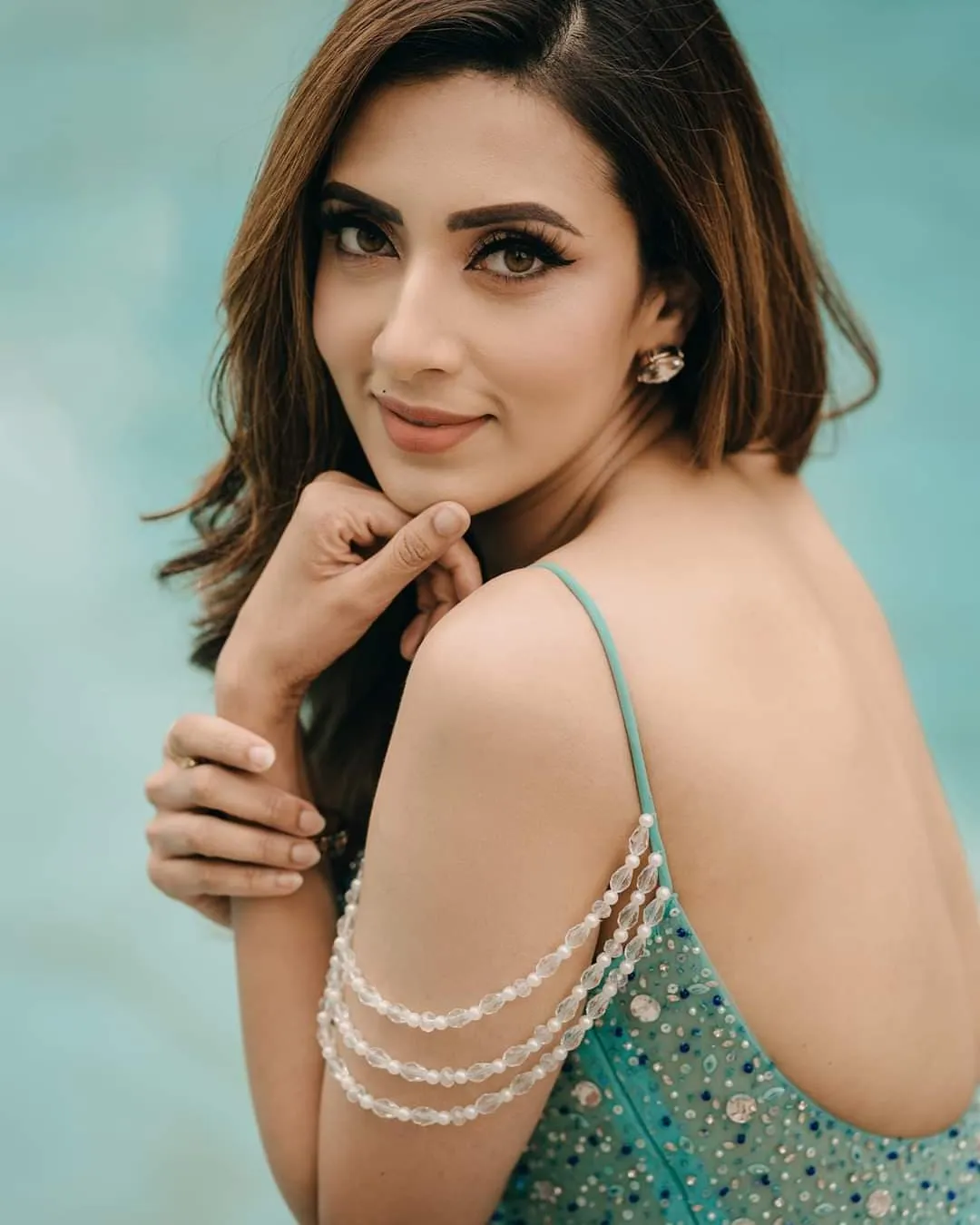Woman in a teal outfit with pearl-draped straps, a hand at her chin, in front of a pale blue backdrop