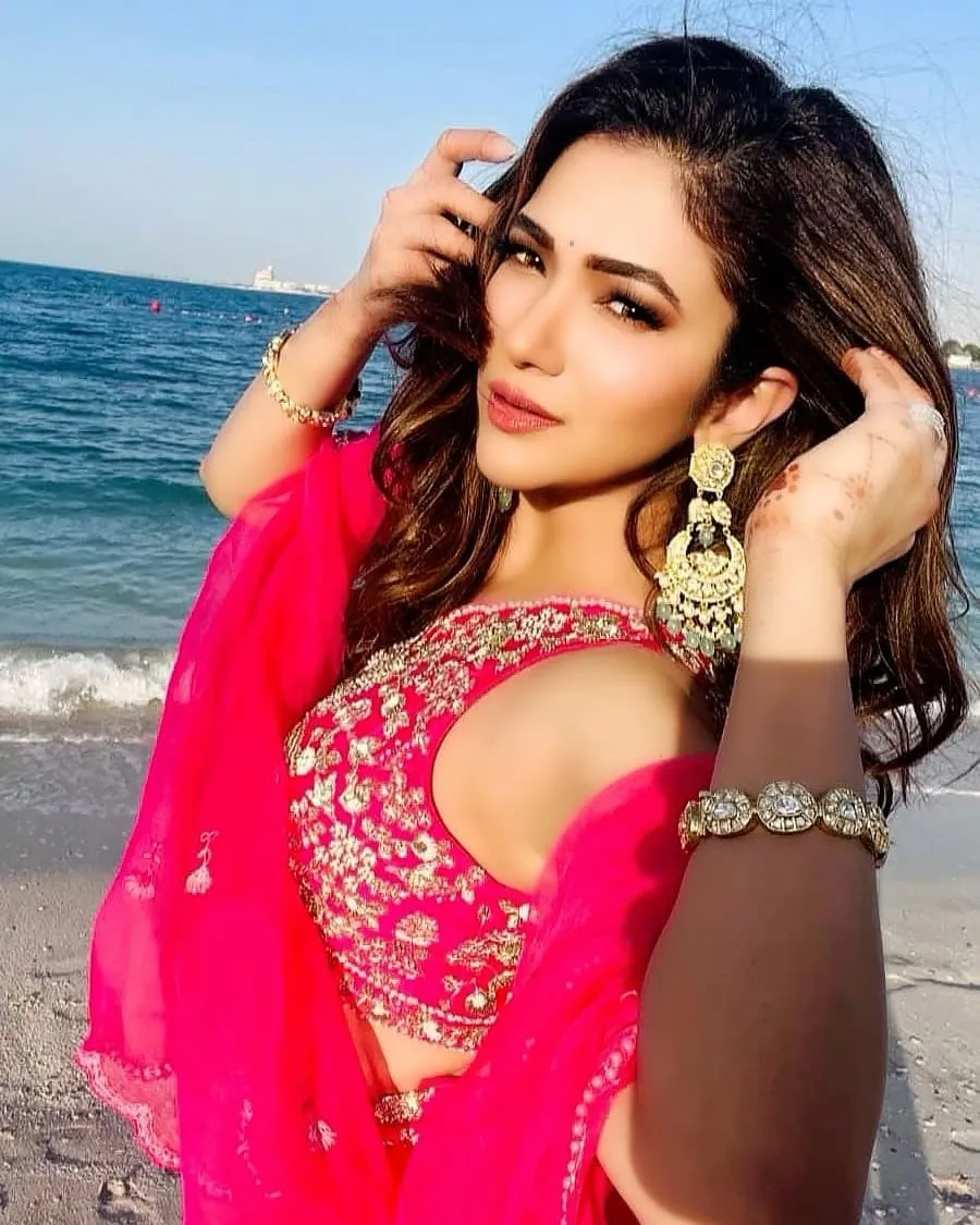 Woman in a pink embellished outfit and gold jewellery with a hand at her ear by the sea