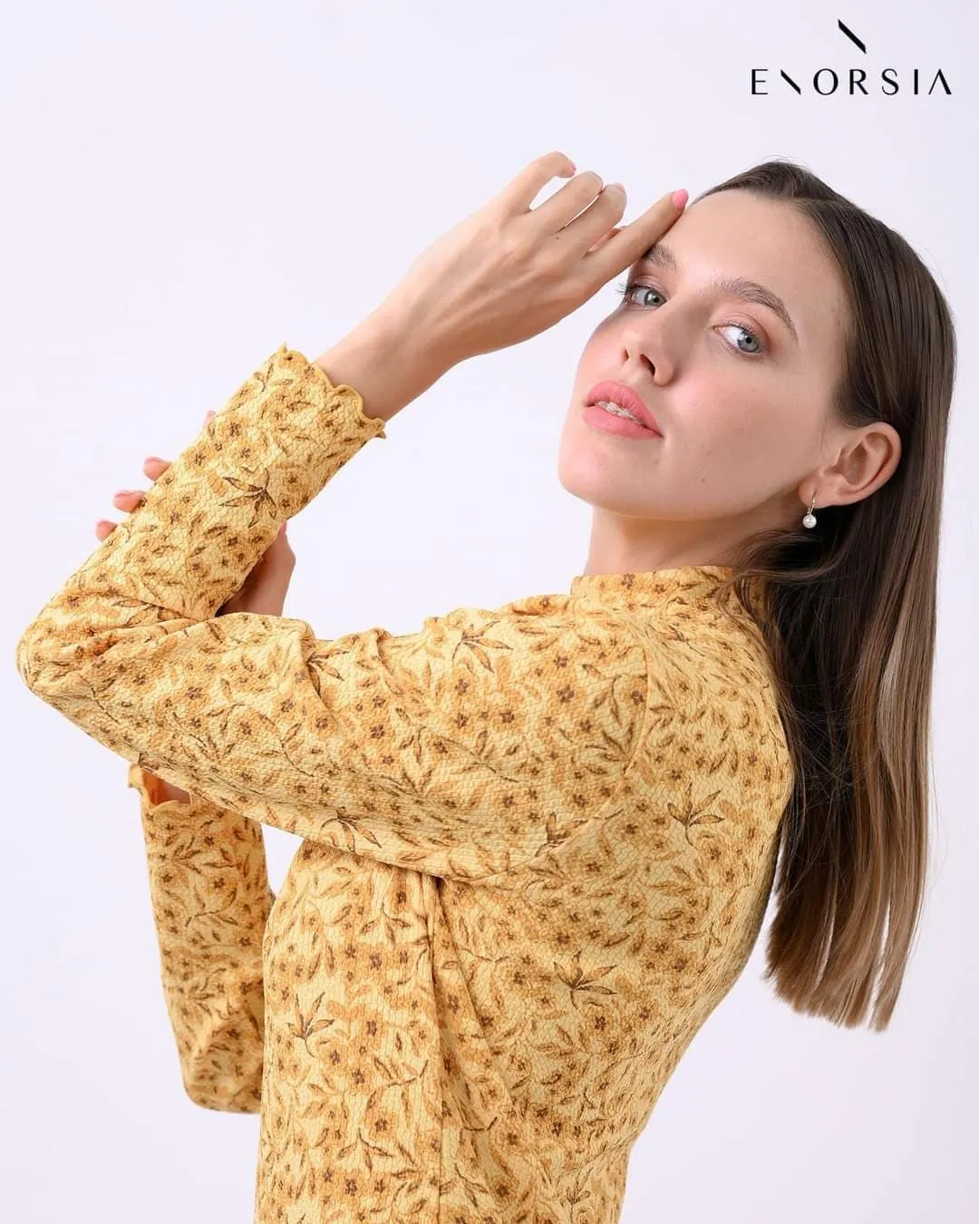 Woman in a mustard-yellow floral top facing the camera with a hand at her forehead against a white backdrop