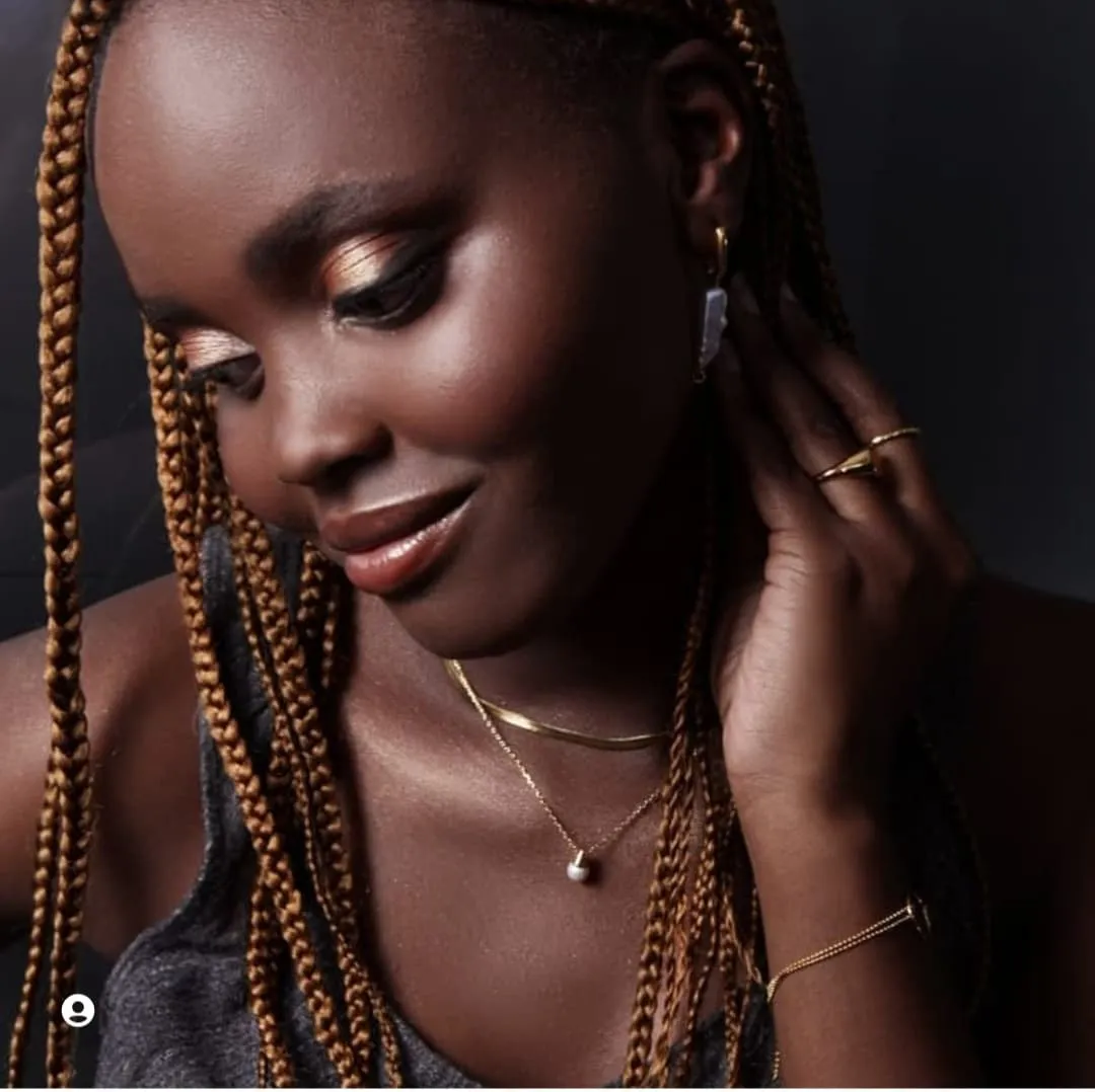 Close-up of a woman with long beaded braids looking down, shimmering gold eyeshadow and a hand at her ear