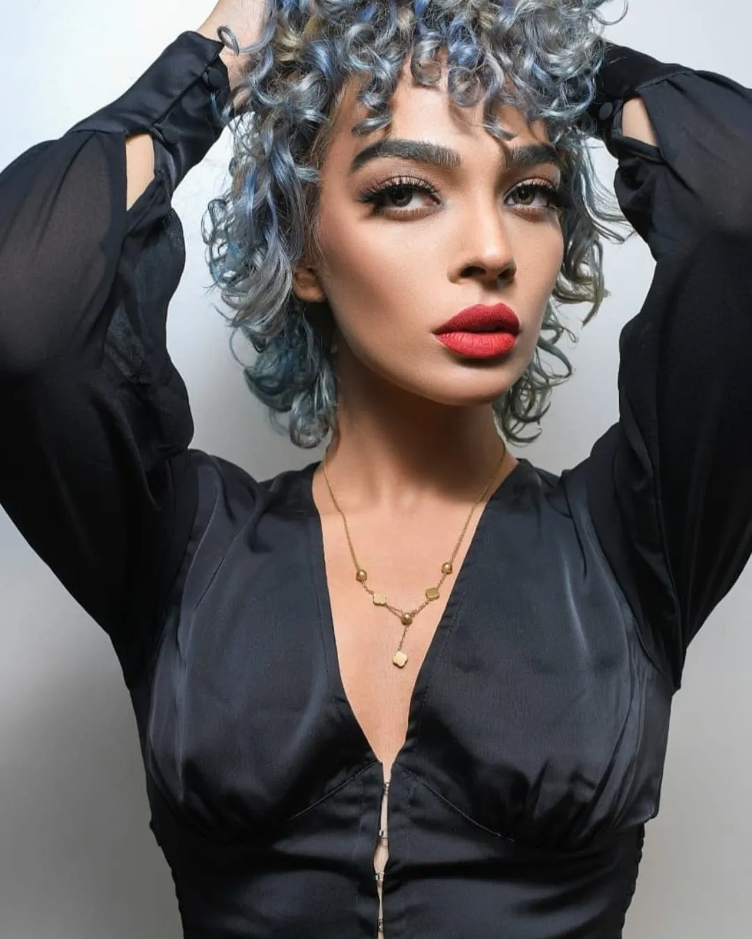 Woman with silver-blue curly hair in a black blouse, both hands in her hair