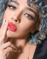 Close-up of a woman with silver-blue curly hair and red lips, a fingertip against her lips