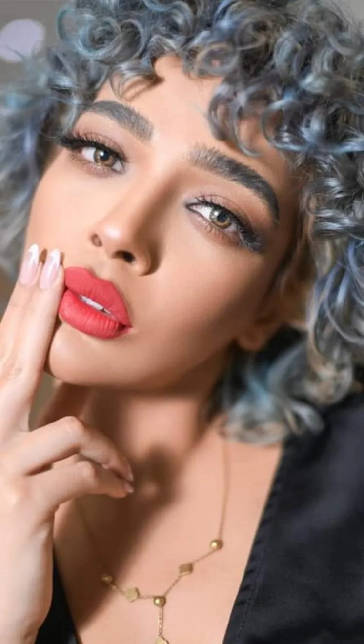 Close-up of a woman with silver-blue curly hair and red lips, a fingertip against her lips