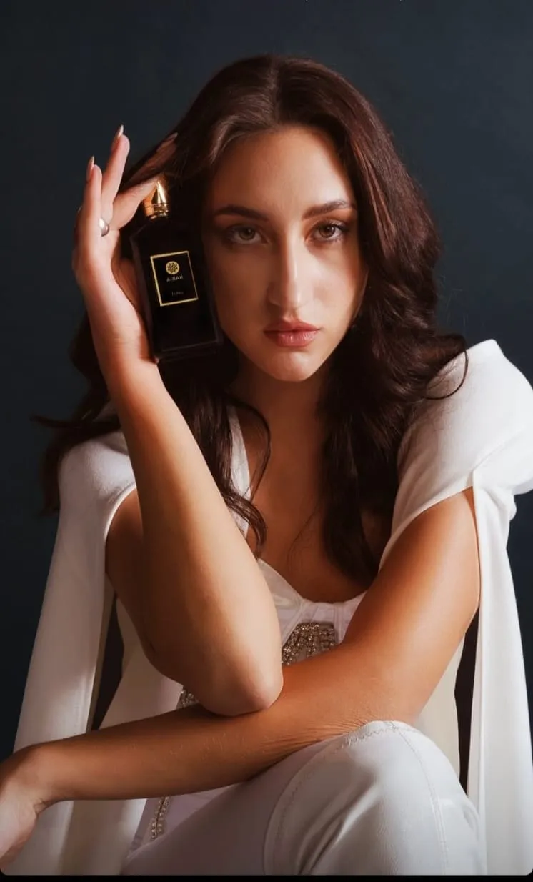 Woman with dark wavy hair in a white jacket against a dark background, a hand at her head