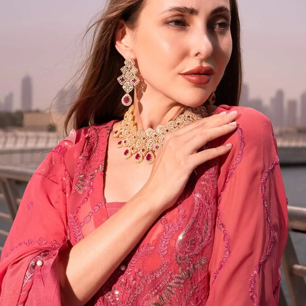 Woman in a pink embroidered outfit with a gold necklace, a hand at her collar, with a city skyline behind