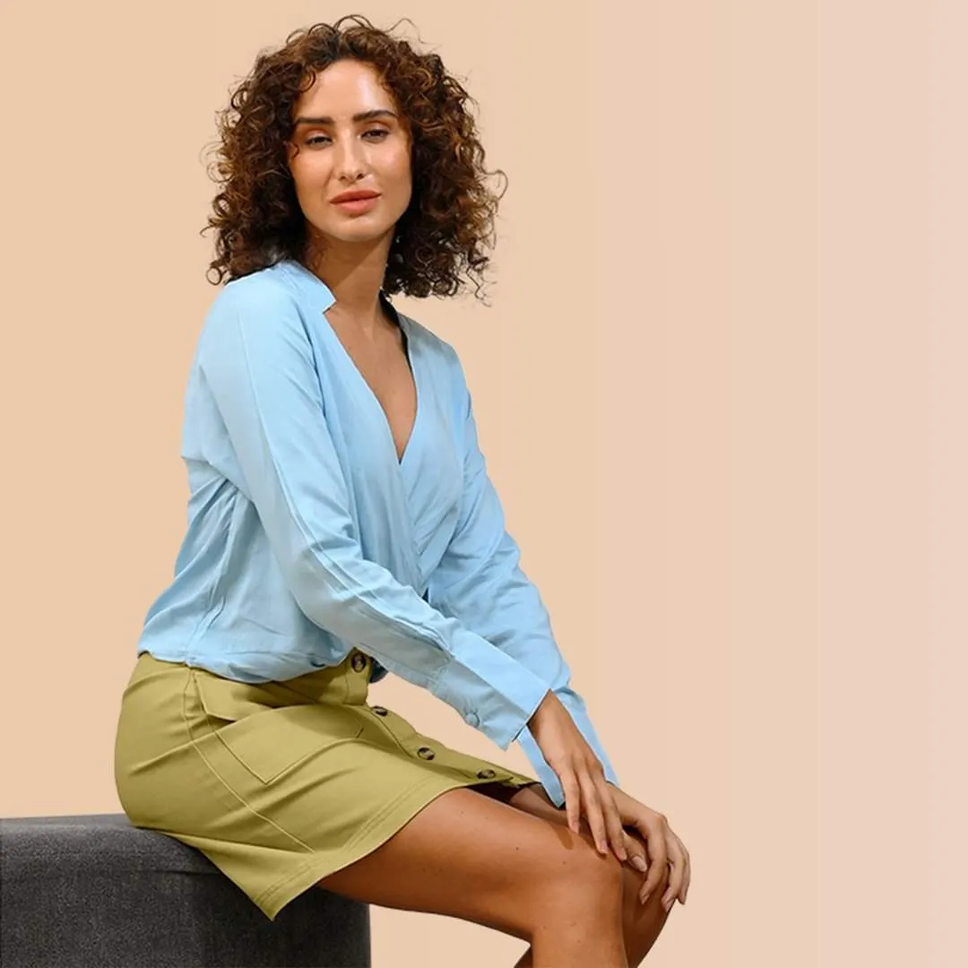 Woman with curly hair in a light-blue shirt and olive skirt seated against a peach backdrop