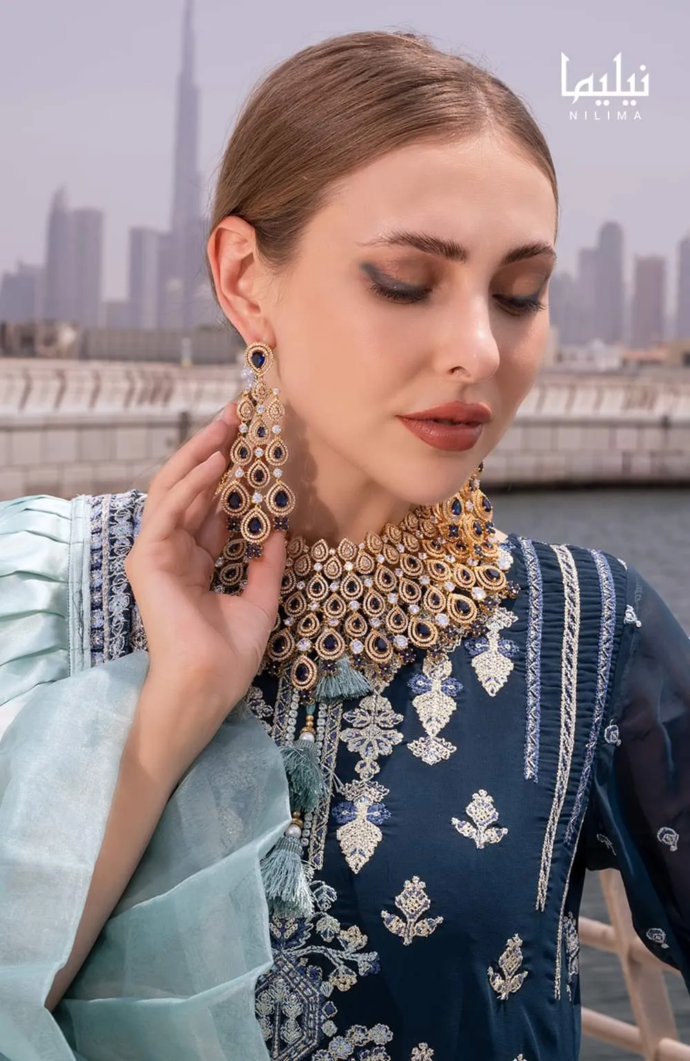 Woman with blonde hair holding both hands near her ears to show large gold and green statement earrings, in a navy embroidered top