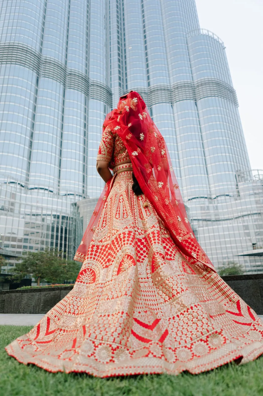 Bride seen from behind in a red and cream embroidered lehenga in front of a tall tower