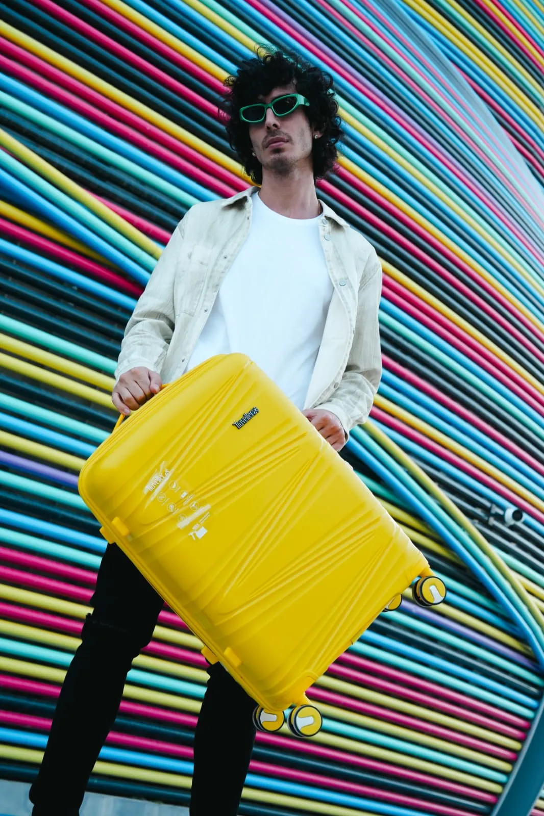 Young man in a white jacket crouching beside a yellow suitcase in front of a rainbow-striped wall