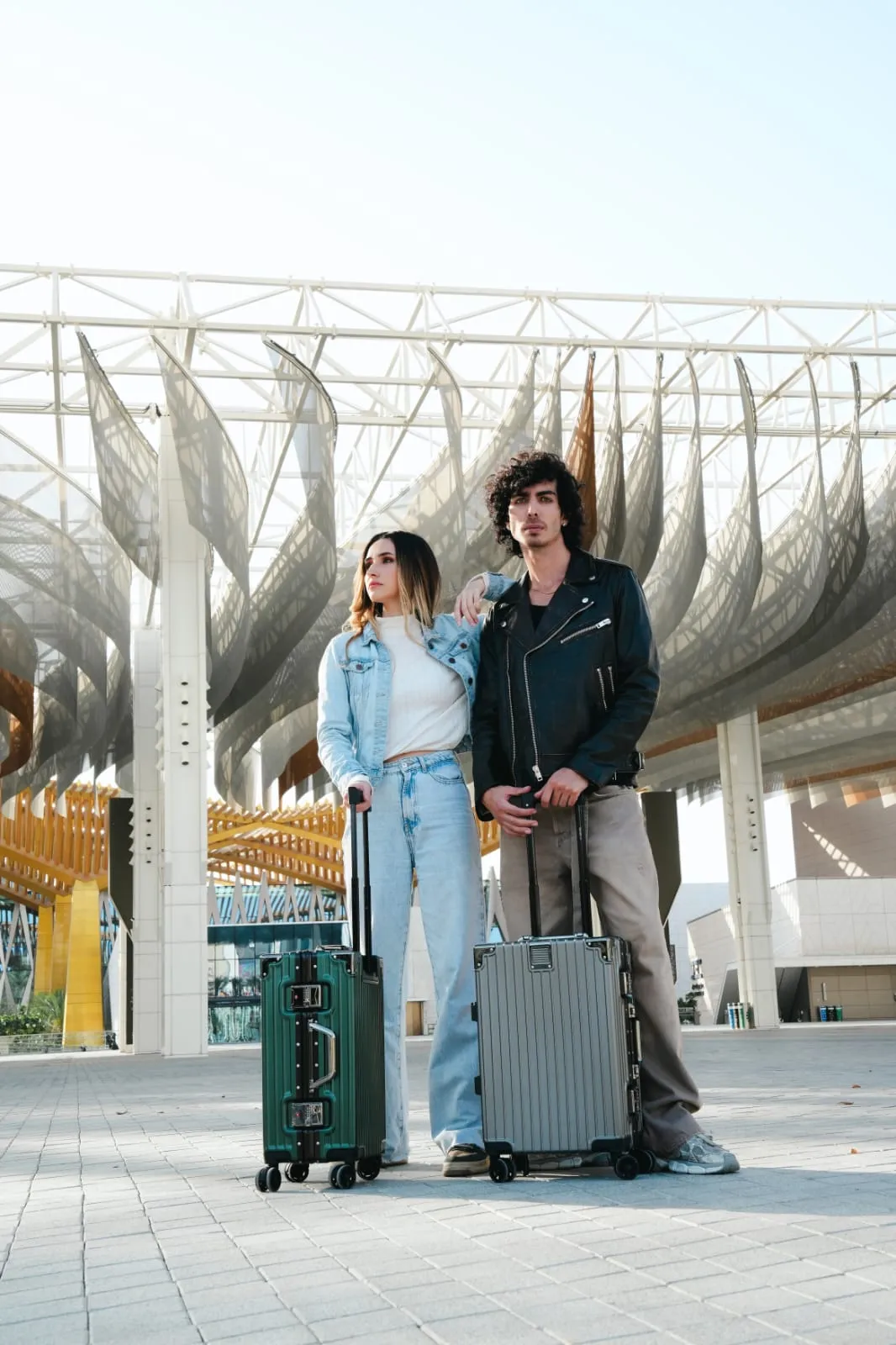 Two people in light and dark outfits standing with suitcases under a canopy-like structure