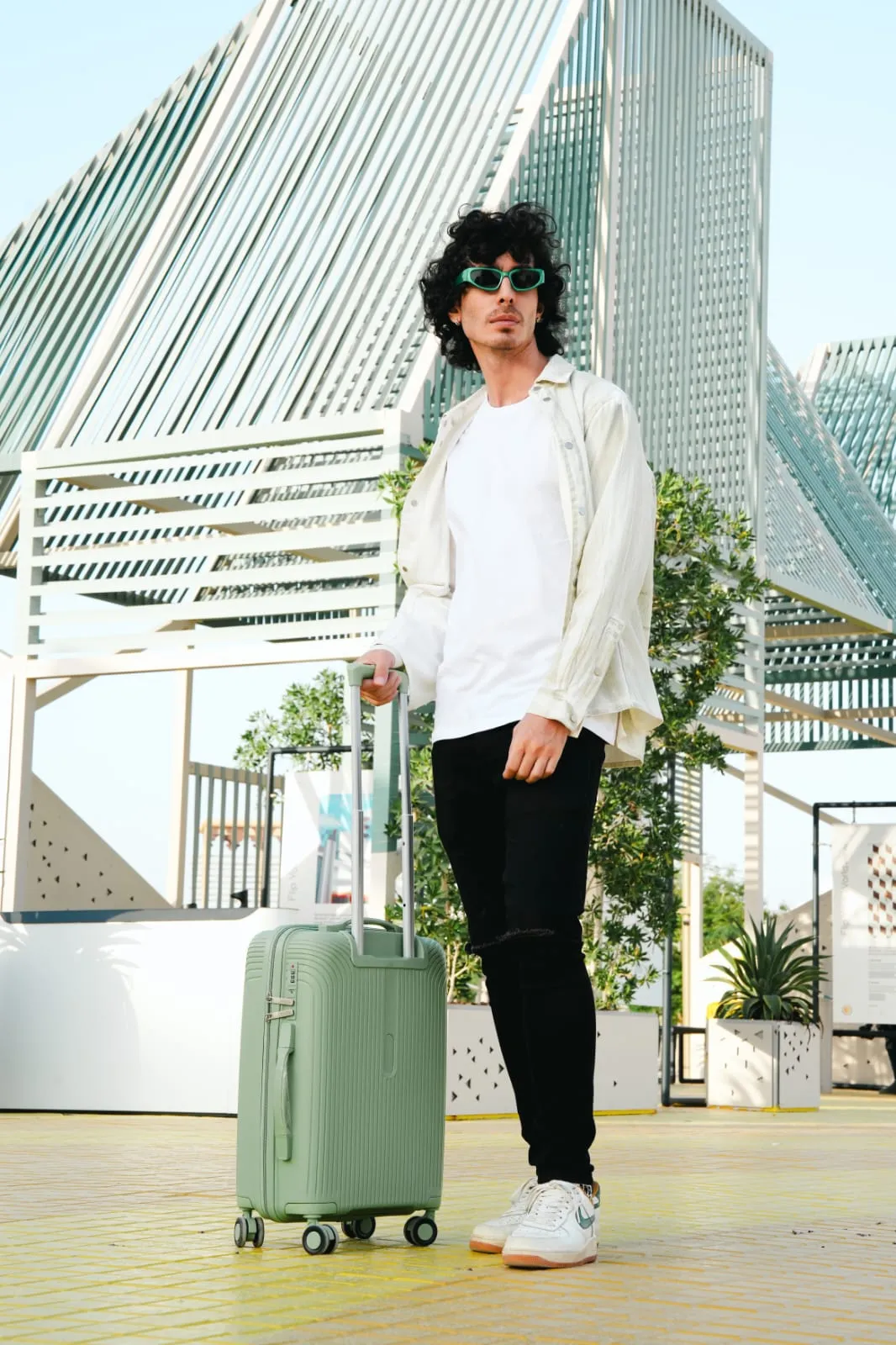 Young man in a cream jacket standing with a green suitcase in front of a glass building
