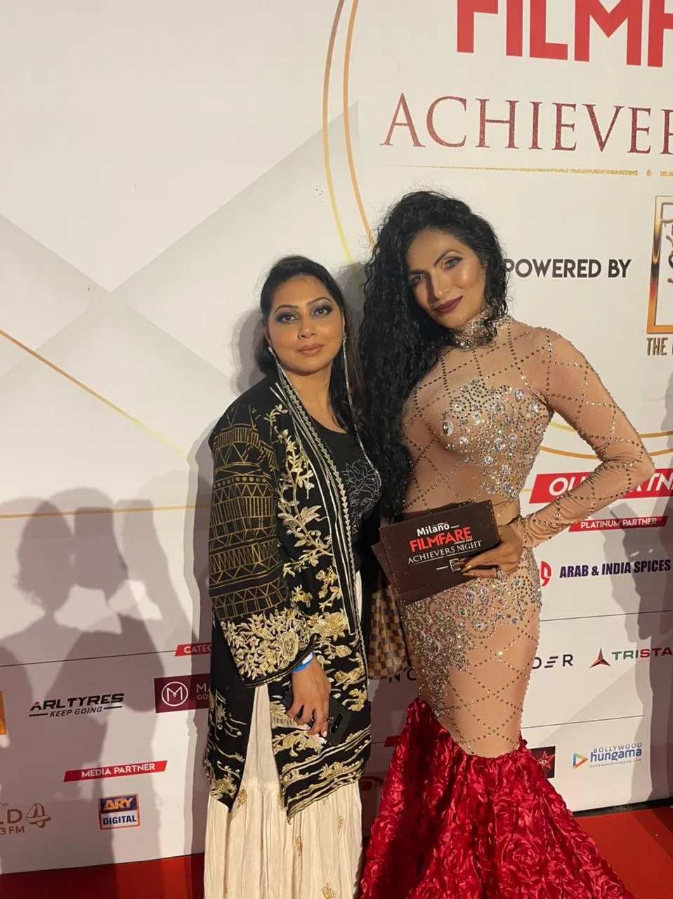 Two women posing in front of an awards-night backdrop, one in a black and gold jacket and one in a sparkly dress with a red train