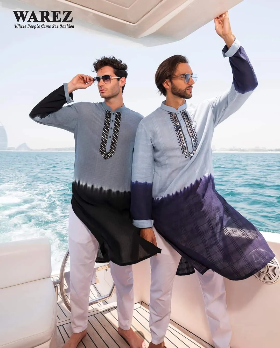 Two men in grey-blue embroidered outfits posing on a yacht with the sea behind
