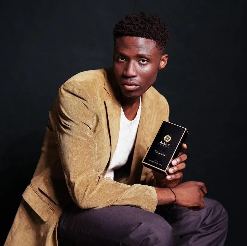 Man in a tan blazer sitting with a black box in his hand against a dark backdrop