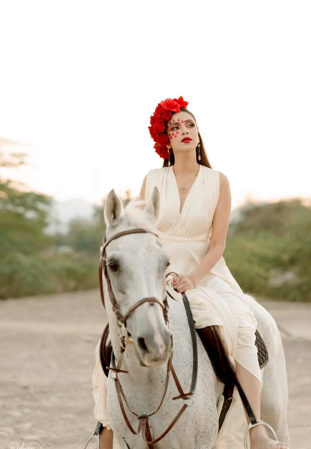 Woman in a cream dress and a red flower headpiece sitting on a white horse in the desert