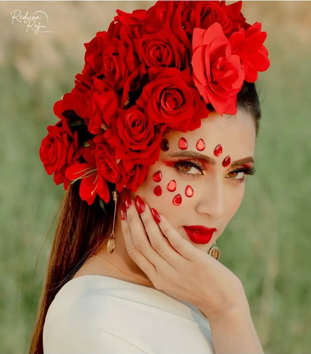 Woman with a headpiece of red roses and red gems on her cheek, a hand at her chin, in front of grass