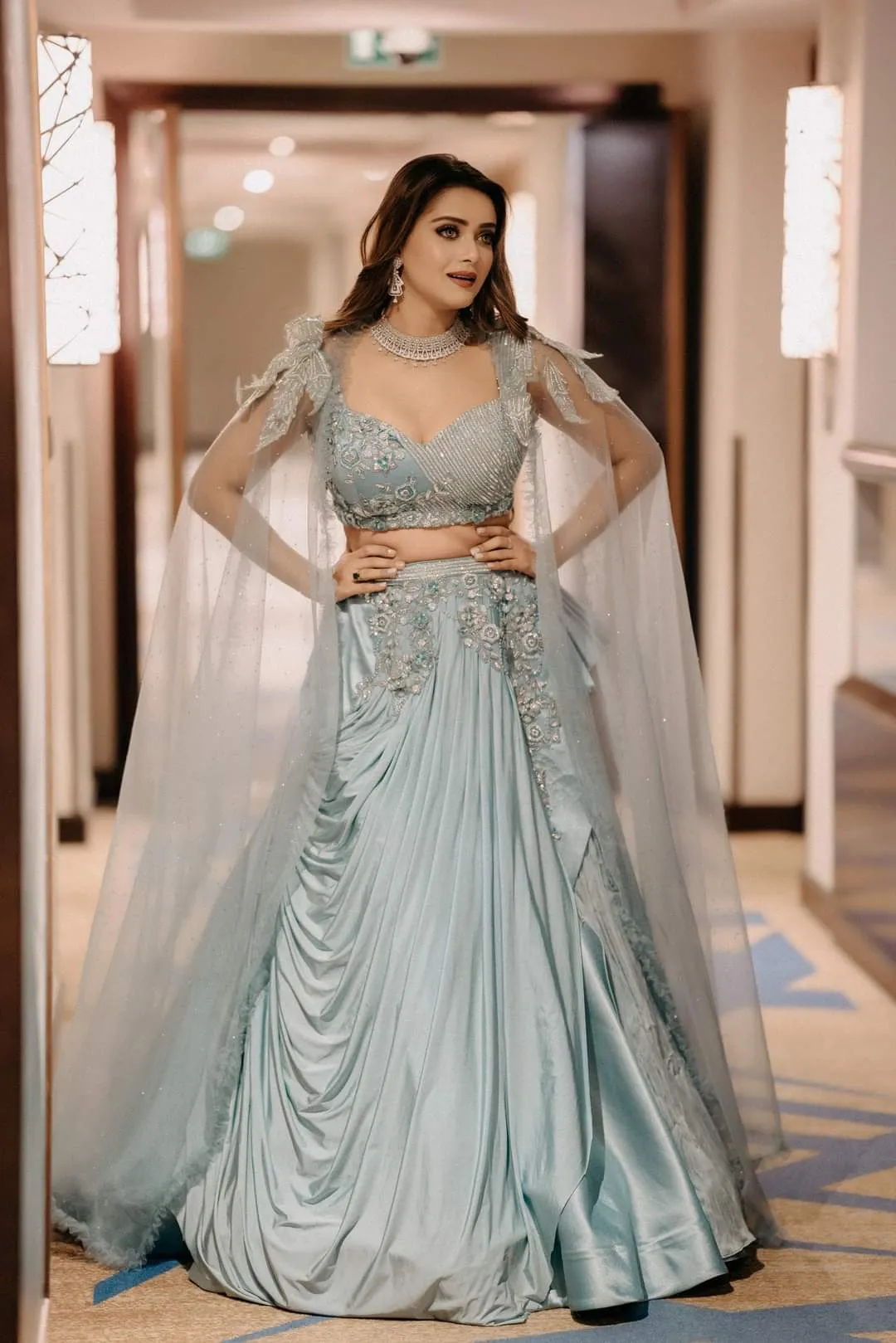 Woman in an embellished sage-green gown with a sheer cape in a hotel corridor
